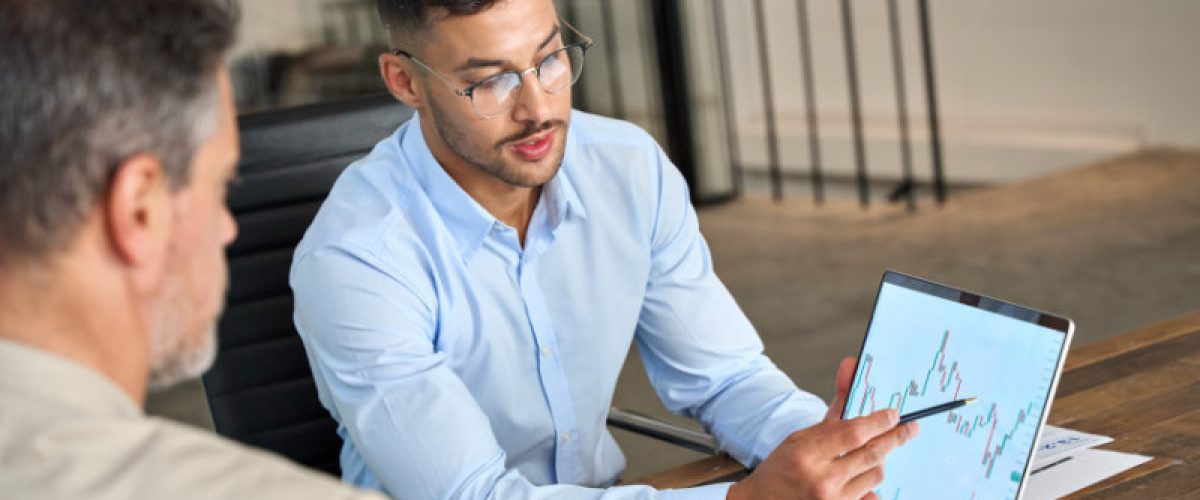Financial advisor explaining invest stock market data consulting investor. Two busy business men analysts doing finance trading analysis pointing at exchange chart on laptop screen working in office.
